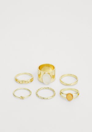 Set di sette anelli d'oro con diversi design: fasce lisce, texture intrecciate e accenti in pietra bianca e arancione.