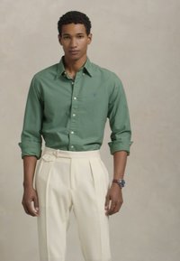 Zelena srajca s gumbi in teksturiranim zaključkom, v kombinaciji z visokimi kremnimi hlačami z gubami in detajlom z buckom na strani.