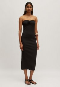 Schwarzes, trägerloses Midikleid mit einer figurbetonten Silhouette und Raffung im Brustbereich. Das Model trägt schwarze flache Sandalen und Holzarmbänder.