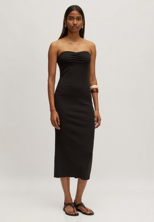 Zwarte, strapless midi-jurk met een getailleerde silhouet en gerimpelde details bij de buste. Model draagt zwarte platte sandalen en houten armbanden.