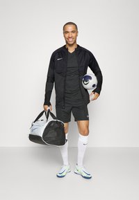 Schwarze Sportjacke mit Reißverschluss, schwarzes T-Shirt, schwarze Shorts, weiße Socken und helle Turnschuhe. Hält einen Fußball und eine Sporttasche.