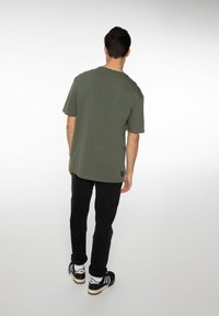 Olijfgroene katoenen t-shirt, ontspannen pasvorm, ronde hals, korte mouwen. Gepaard met zwarte broeken en zwarte sneakers met witte accenten.