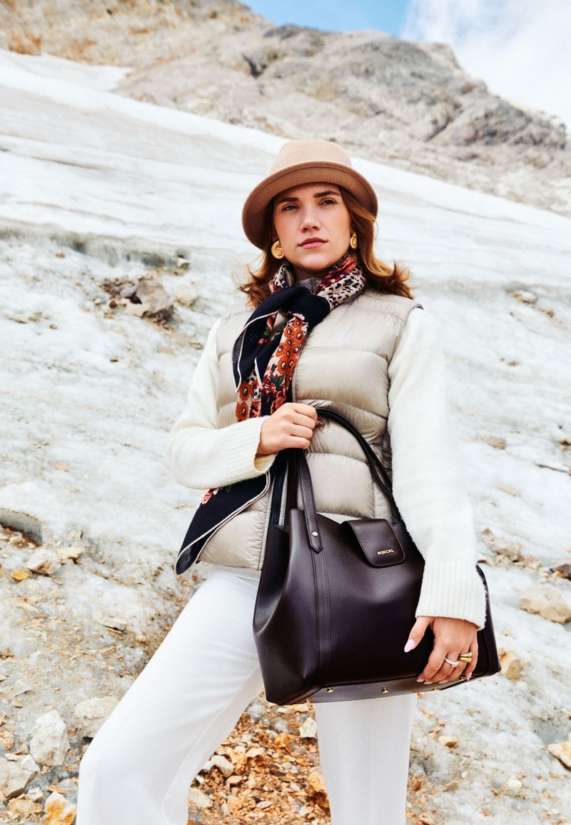 Large dark brown leather handbag with minimal hardware, worn by a model in a cream sweater and beige vest, styled with a patterned scarf.
