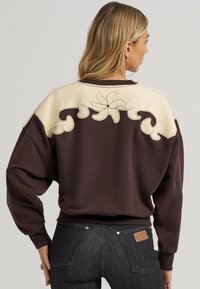 Καφέ φούτερ με κρεμ πάνελ και λουλουδάτη κεντητική διακόσμηση στην πλάτη; ραμμένα μανίκια και τελείωμα; χαλαρή εφαρμογή, φορεμένο με σκούρα τζιν.