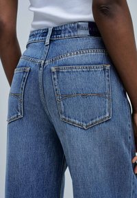 Dark-skinned person wearing light blue denim jeans with back pockets and a white ribbed top, shown from the waist down to mid-thigh.