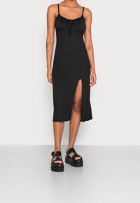 Mujer vestida con un vestido midi sin mangas negro con abertura frontal y sandalias de plataforma negras, de pie contra un fondo blanco liso.