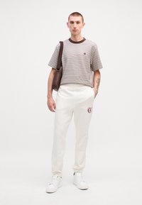 Pasiasta brązowo-biała koszula z krótkim rękawem, kremowe dresowe spodnie z logo, białe sneakersy, trzymając brązową torbę na ramię, stojąc na tle białym.