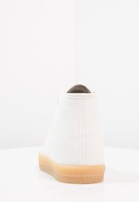 Biały płócienny sneaker z jasnobrązową gumową podeszwą, pokazany z tyłu na białej powierzchni z jednolitym białym tłem.