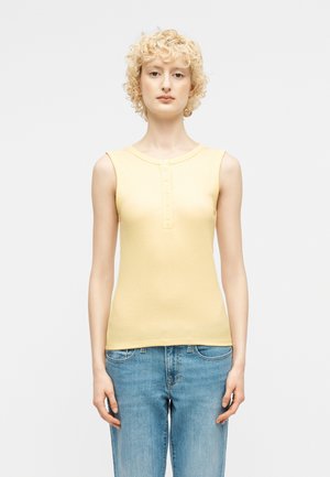 Personne aux cheveux blonds bouclés courts portant un haut sans manches jaune pâle boutonné et un jean bleu, debout devant un fond uni.