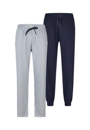 Oberkörperfreier Mann mit detailliertem Vollarm-Tattoo am rechten Arm, trägt grau-weiß gestreifte Hosen mit vertikalen Streifen, steht barfuß, schaut nach rechts und lächelt.