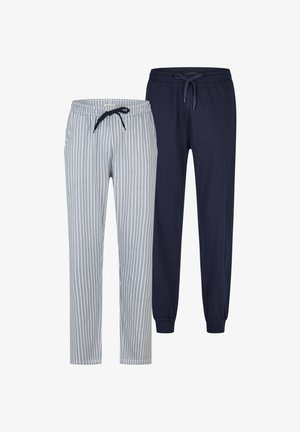 Oberkörperfreier Mann mit detailliertem Vollarm-Tattoo am rechten Arm, trägt grau-weiß gestreifte Hosen mit vertikalen Streifen, steht barfuß, schaut nach rechts und lächelt.