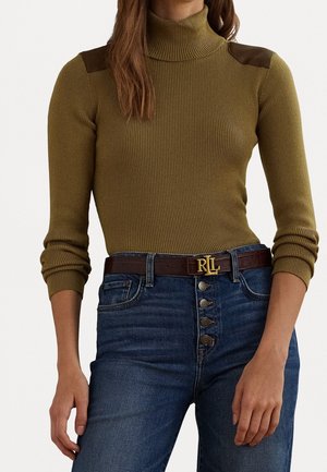 Femme portant un pull à col roulé côtelé olive avec des patchs en cuir aux épaules et un jean bleu taille haute avec une ceinture marron.