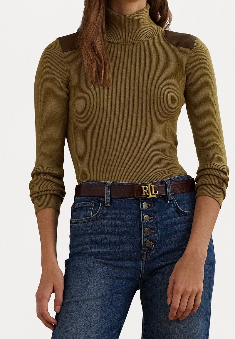 Femme portant un pull à col roulé côtelé olive avec des patchs en cuir aux épaules et un jean bleu taille haute avec une ceinture marron.