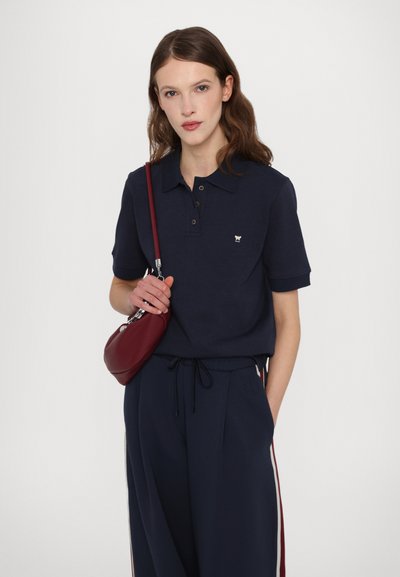 Marine polo-skjorte med korte ermer, tre-knapps placket og liten brodert logo. Kombinert med marineblå bukser og en bordeaux håndveske.
