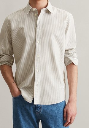 Mann trägt beige-weiß gestreiftes, geknöpftes Hemd mit hochgekrempelten Ärmeln und blaue Jeans, eine Hand in der Tasche.