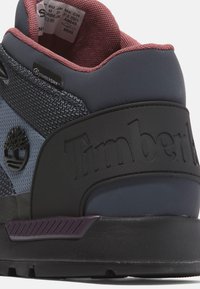 Zapatilla negra y azul marino con un collar acolchado, tejido texturizado y logo de Timberland. Presenta un acento morado en contraste en la suela.