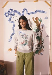 Hall halli pusa, millel on värvikas loodusteemaline trükk, sobitatud roheliste volditud pükstega. Taustal on kunstipärased dekoratsioonid.