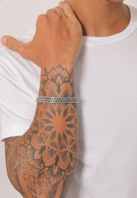 Silverfärgad flätad armband på ett handled med intrikata blommigt tatueringar. Armbandet har en texturerad, flätad design mot en ljus bakgrund.