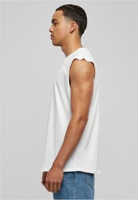 Jeune homme aux cheveux courts portant un t-shirt sans manches blanc et un jean bleu, se tenant de profil contre un fond gris uni.