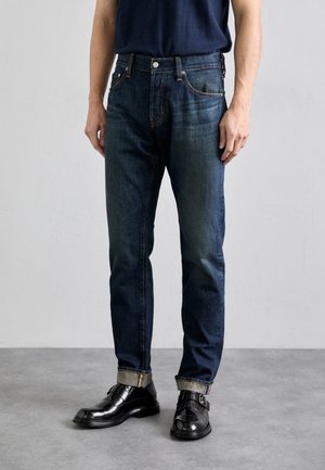 Mörkblå denimjeans med slim fit, med orange sömmar, fem fickor och uppvikta ben, kombinerat med glänsande svarta dresskor.