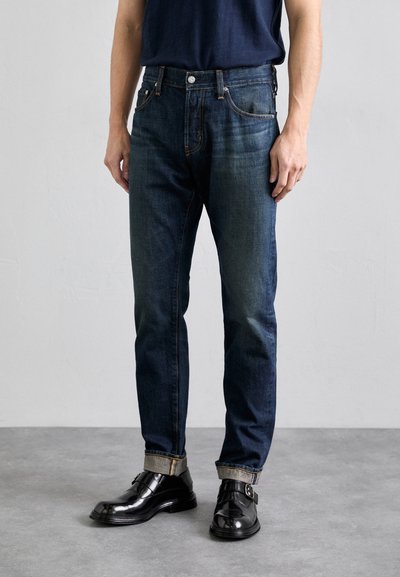 Tmavě modré džíny s úzkým střihem, zdobené oranžovým prošíváním, pěti kapsami a ohrnutým lemem, v kombinaci s lesklými černými společenskými botami.