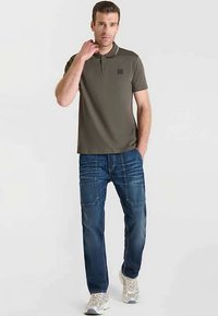 Olijfgroene polo met kraag en een klein logo, gecombineerd met lichtblauwe spijkerbroek voorzien van voorzakken en lichte vervaging. Casual schoenen.