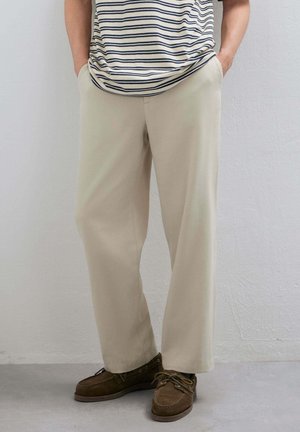 Persona che indossa pantaloni beige a gamba dritta, mani in tasca, abbinati a mocassini marroni con lacci e una camicia bianca e blu navy a righe.