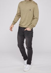 Maglione a collo alto beige in maglia con un piccolo logo nero sul petto, abbinato a jeans skinny scuri e sneaker bianche.