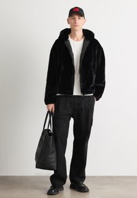 Black faux fur zip-up jacket with hood, white t-shirt, black cargo pants, and a black tote bag. Model wears a black cap with red logo.
