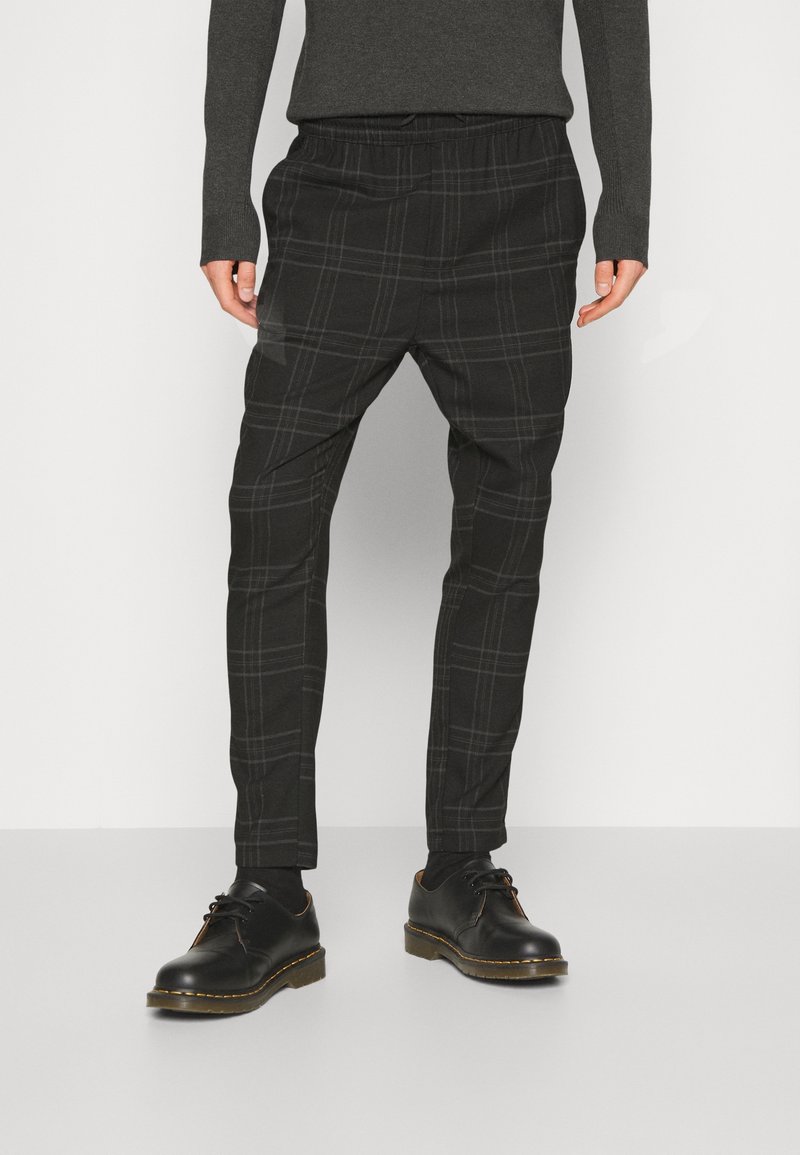 Zwarte geruite broek met een elastische tailleband, taps toelopende pijpen en een gestructureerde stof, gecombineerd met zwarte veterschoenen.