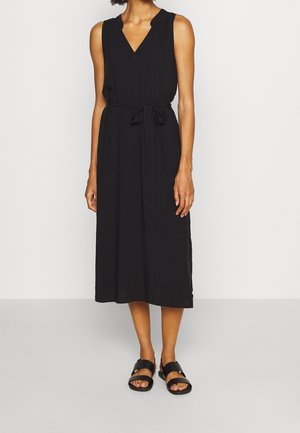 Nainen yllään musta hihaton midimekko, jossa V-pääntie ja sidottu vyötärönauha, yhdistettynä mustiin avokkaisiin sandaaleihin yksinkertaisella taustalla.