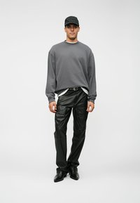 Giovane uomo che indossa un berretto nero, una felpa grigia oversize, pantaloni cargo in pelle nera e scarpe nere appuntite, in piedi davanti a uno sfondo semplice.