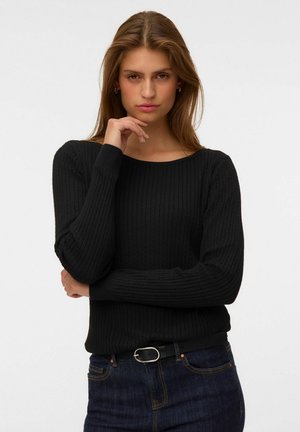 Jeune femme aux longs cheveux bruns porte un pull noir côtelé et un jean bleu foncé, posant avec une main près du menton sur un fond uni.