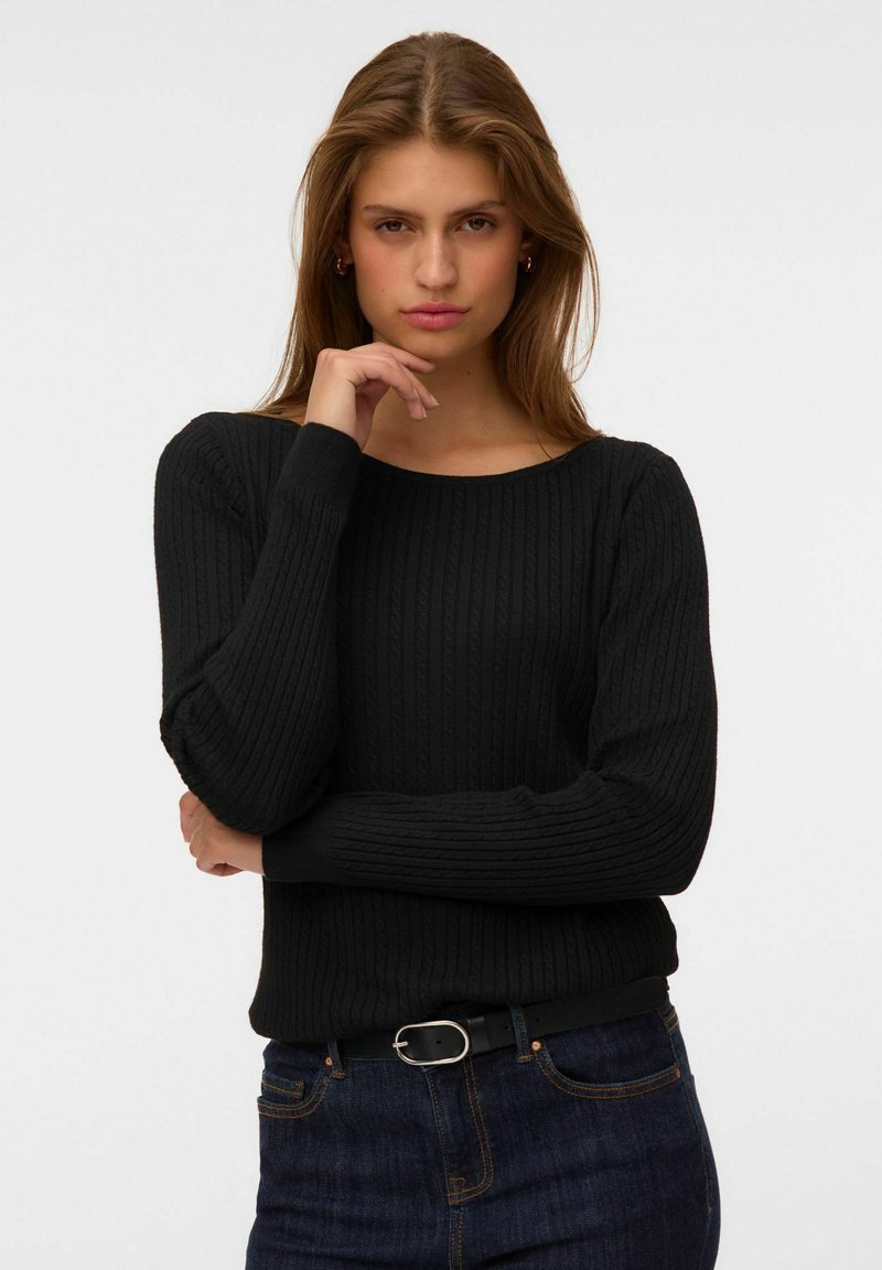 Jeune femme aux longs cheveux bruns porte un pull noir côtelé et un jean bleu foncé, posant avec une main près du menton sur un fond uni.