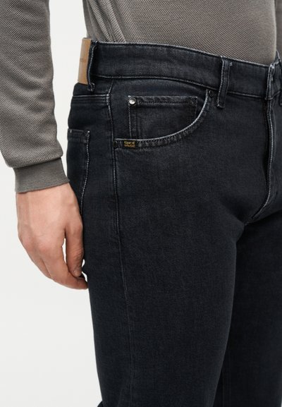 Άνδρας που φοράει σκούρο μαύρο τζιν και γκρι υφασμάτινο μπλουζάκι με μακριά μανίκια, δείχνοντας την μπροστινή τσέπη και το χέρι που ακουμπάει πλάι στο μηρό.