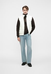 Jaunas vyras stovi vilkėdamas baltą siūtuoti liemenę ant tamsaus užtrauktuku užsegamo megztinio, šviesiai mėlynas džinsus ir juodas loaferius ant balto fono.