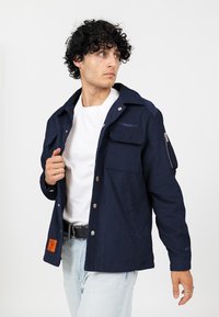 Marineblauwe corduroyjas met metalen knopen, twee voorzakken en een verstelbare zak op de mouw. Draagt over een wit T-shirt.