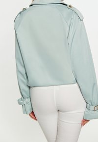 Trench-coat bleu clair raccourci avec boutons aux épaules, poignets retroussés et texture lisse, associé à un pantalon blanc ajusté.