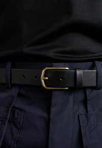 Schwarzer Ledergürtel mit einer goldenen rechteckigen Schnalle, getragen über blauen Hosen. Das Leder hat eine glatte Textur und verfügt über gestanzte Löcher zur Anpassung.