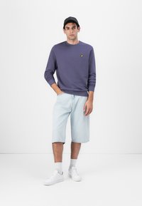 Felpa di cotone viola con logo giallo, pantaloni shorts in denim chiaro e sneakers bianche, con un design comodo e colletto a girocollo.