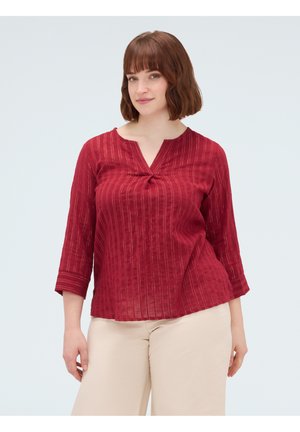 Donna con capelli castani fino alle spalle che indossa una camicetta rossa a righe con maniche lunghe e pantaloni beige, in piedi davanti a uno sfondo chiaro.