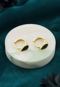 Orecchini a cerchio in tono dorato con inserti ovali neri. Sono esposti su una superficie rotonda di pietra chiara.