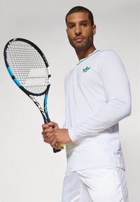 Fehér, hosszú ujjú teniszpóló zöld kiegészítőkkel és adidas logóval, párosítva egy fekete és kék keretű teniszütővel.