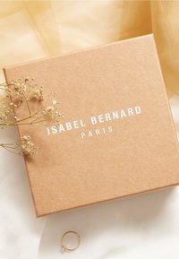 Brown kraft box with white text "ISABEL BERNARD PARIS," alongside a small gold ring and dried flowers, on a light fabric background.