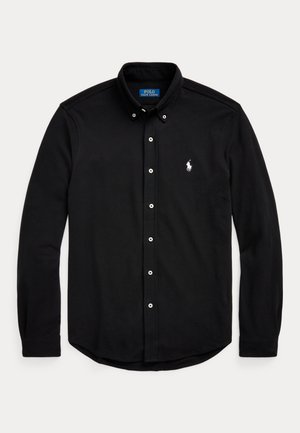 Un homme porte une chemise noire boutonnée, un pantalon noir, une ceinture, une montre-bracelet et des chaussures noires sur un fond de studio beige.