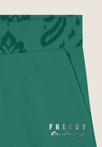 Pantaloni sportivi verdi con una cintura testurizzata che presenta un motivo verde scuro. Include una tasca laterale e dettagli in logo metallico argento.