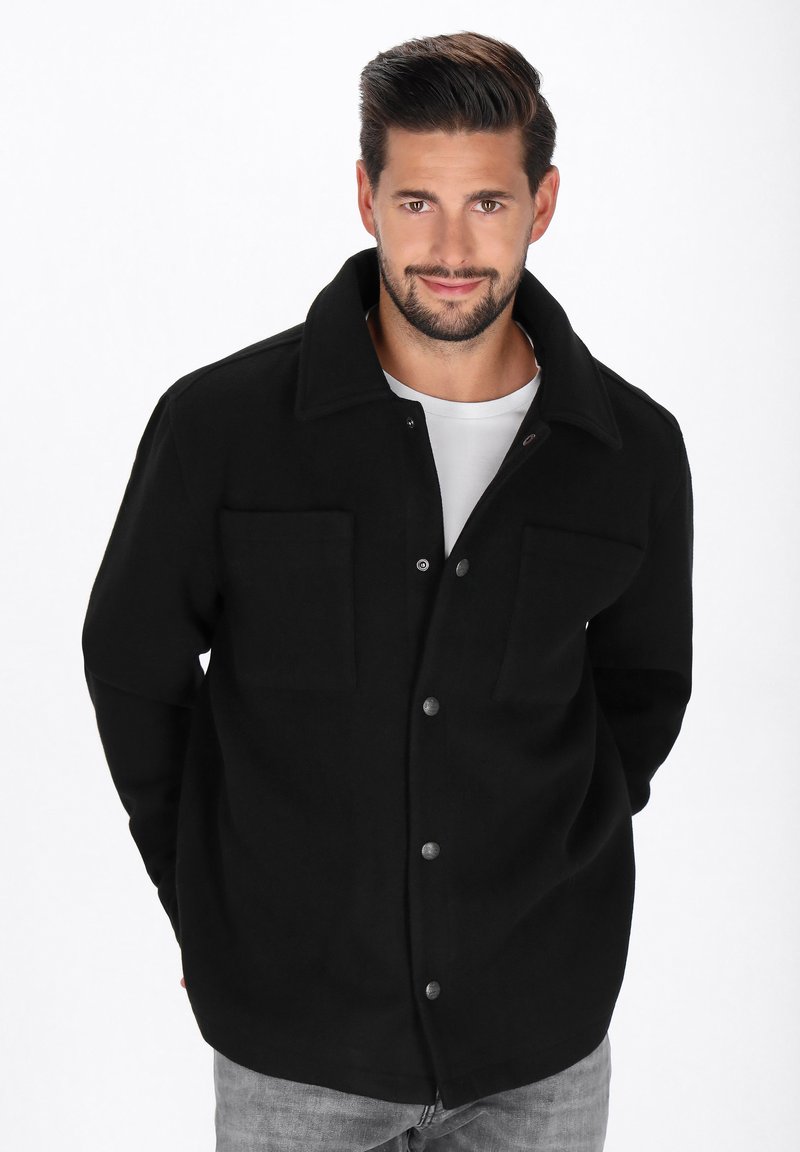 Mann, der eine schwarze, knopfverdeckte Jacke über einem weißen Hemd und grauen Jeans trägt, mit den Händen hinter dem Rücken stehend und in die Kamera lächelnd.