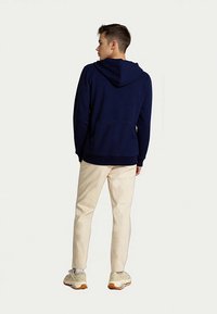 Mladý muž stojící a dívající se do strany, oblečený v tmavě modré mikině s kapucí, béžových kalhotách, bílých ponožkách a béžových teniskách.