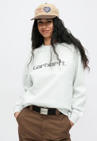 Jasnoszara bluza z nadrukiem "carhartt" w ciemnoszarym kolorze, zestawiona z brązowymi spodniami i beżową czapką z kolorowym napisem oraz grafiką serca.