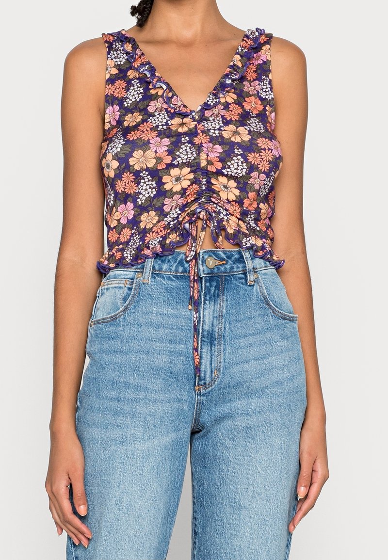 Top court sans manches violet à motif floral avec un volant sur les bords et un cordon à l'avant, associé à un jean taille haute bleu clair.